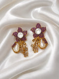 A pair of ornate gold and purple floral earrings with white pearl accents, displayed on a draped white satin fabric background.