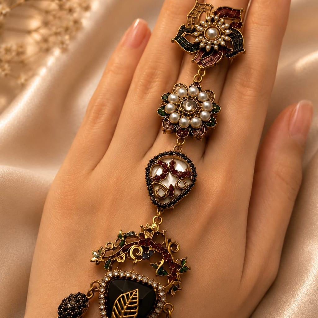 An ornate gold-toned ring with multiple dangling floral and leaf motifs, set with pearls, dark green enamel, and red stones, worn on a hand against a soft beige satin background.