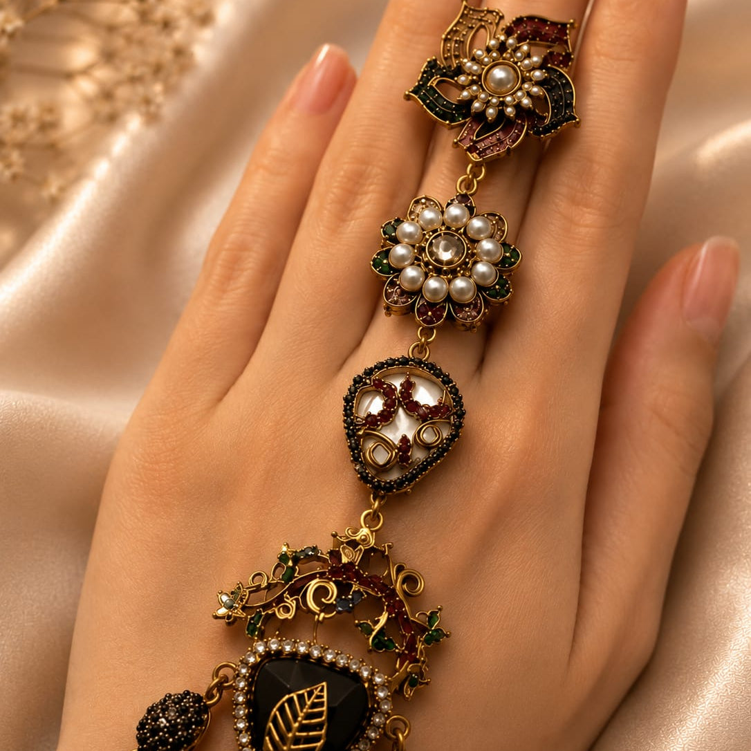 An ornate gold-toned ring with multiple dangling floral and leaf motifs, set with pearls, dark green enamel, and red stones, worn on a hand against a soft beige satin background.