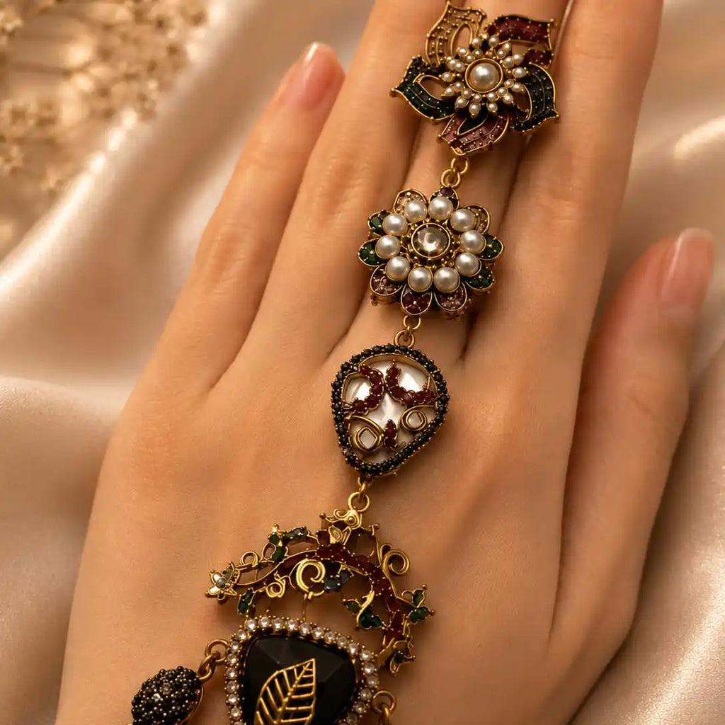 An ornate gold-toned ring with multiple dangling floral and leaf motifs, set with pearls, dark green enamel, and red stones, worn on a hand against a soft beige satin background.